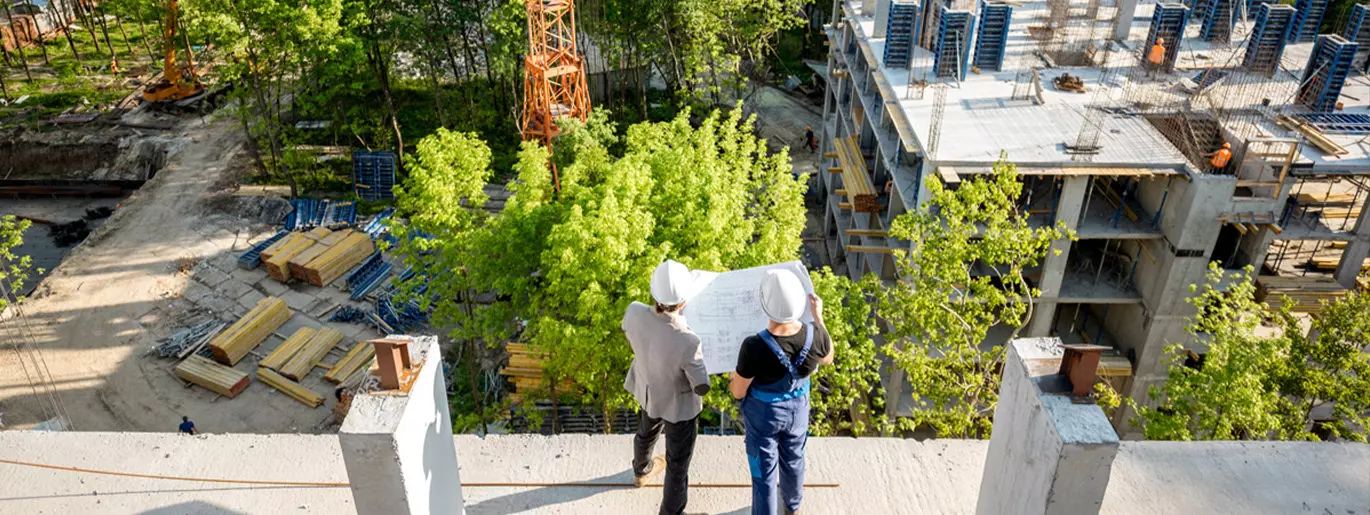 Kaksi rakennustyöläistä seisoo katolla katsoen rakennuspiirustuksia ja alhaalla aukeavaa rakennustyömaata 